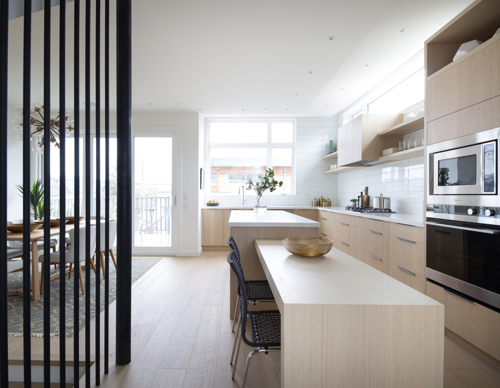 Scandinavian l-shaped kitchen/diner in Vancouver with flat-panel cabinets, light wood cabinets, white splashback, stainless steel appliances, light hardwood flooring, an island, beige floors and white worktops.