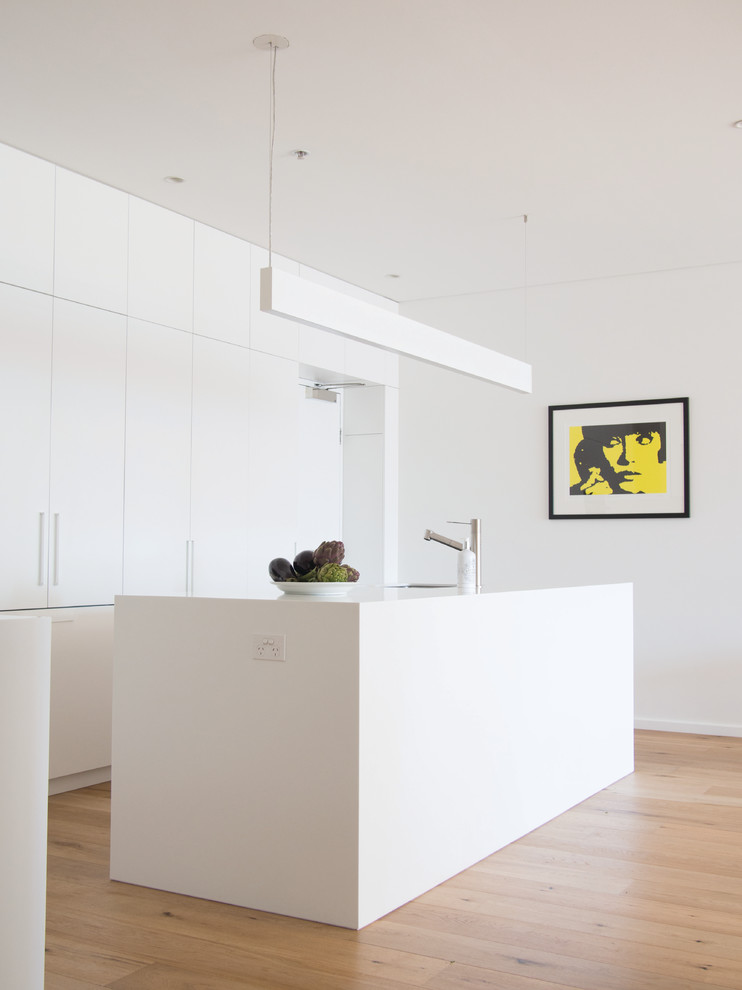 Cette image montre une cuisine encastrable minimaliste en L avec un placard à porte plane, des portes de placard blanches, un sol en bois brun, îlot, un sol marron et un plan de travail blanc.