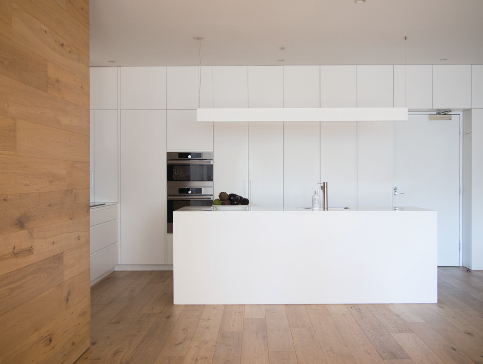Moderne Küche in L-Form mit flächenbündigen Schrankfronten, weißen Schränken, Küchengeräten aus Edelstahl, braunem Holzboden, Kücheninsel, beigem Boden und weißer Arbeitsplatte