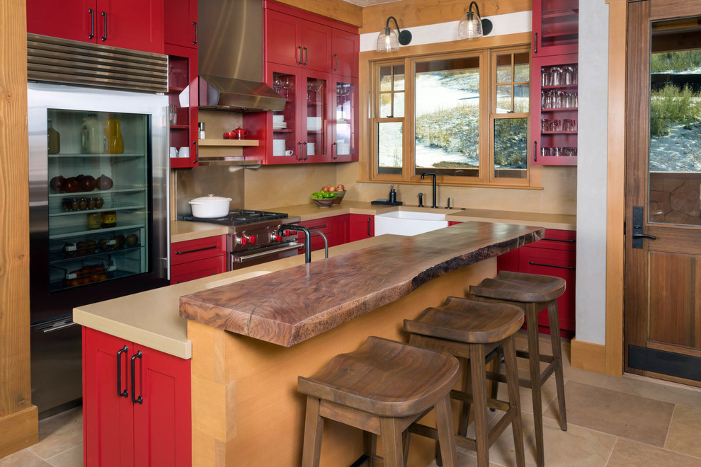 Cette image montre une cuisine chalet avec un évier de ferme, un placard à porte vitrée, des portes de placard rouges, une crédence beige, un électroménager en acier inoxydable et îlot.