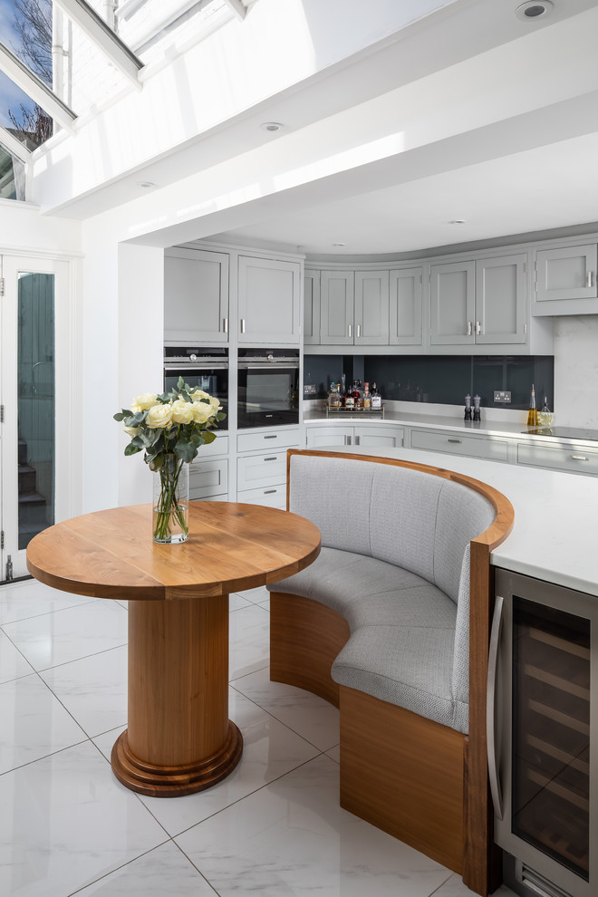 Идея дизайна: угловая кухня в стиле неоклассика (современная классика) с обеденным столом, фасадами в стиле шейкер, серыми фасадами, фартуком из стекла, островом, белым полом и белой столешницей