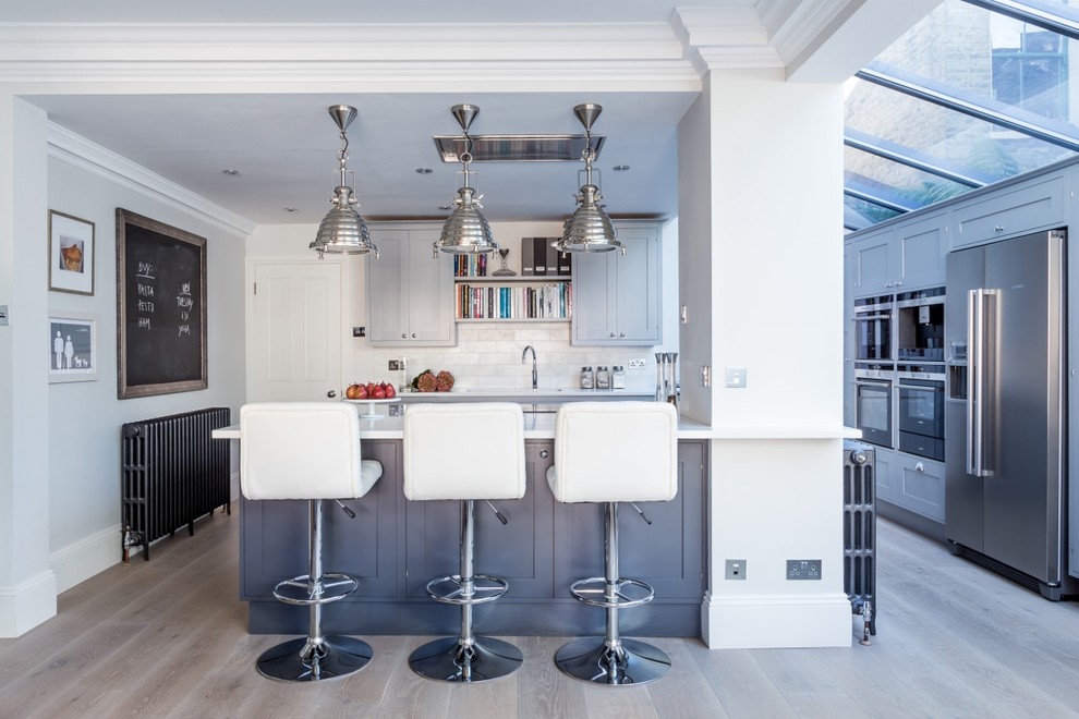 Inspiration for a classic l-shaped kitchen in London with shaker cabinets, grey cabinets, white splashback, metro tiled splashback, stainless steel appliances, light hardwood flooring and a breakfast bar.