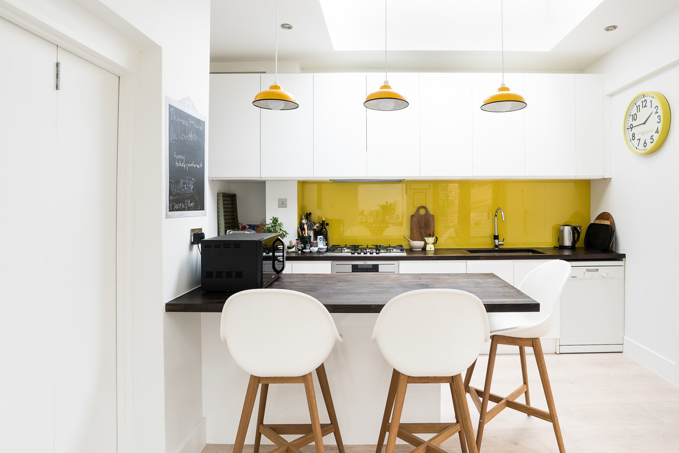 This is an example of a contemporary galley kitchen in London with a submerged sink, flat-panel cabinets, white cabinets, wood worktops, yellow splashback, glass sheet splashback, white appliances, a breakfast bar, beige floors and brown worktops.