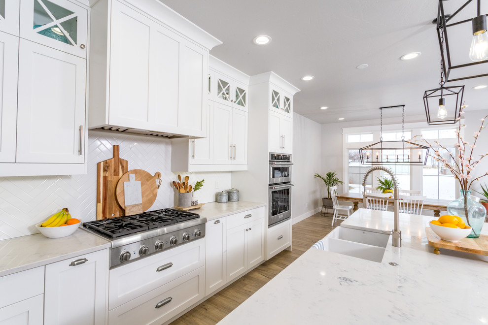 Photo of a large beach style l-shaped kitchen/diner in San Francisco with a belfast sink, shaker cabinets, white cabinets, engineered stone countertops, white splashback, cement tile splashback, stainless steel appliances, light hardwood flooring, an island, beige floors and white worktops.