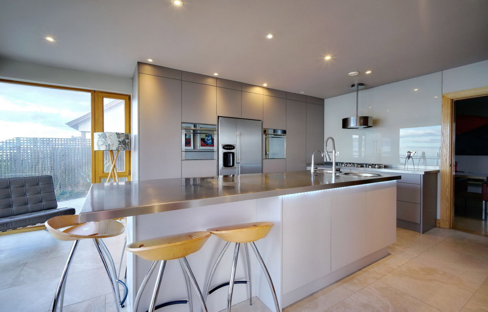 Large modern l-shaped enclosed kitchen in Belfast with a built-in sink, flat-panel cabinets, beige cabinets, stainless steel worktops, beige splashback, glass sheet splashback, stainless steel appliances, ceramic flooring, an island, beige floors and grey worktops.