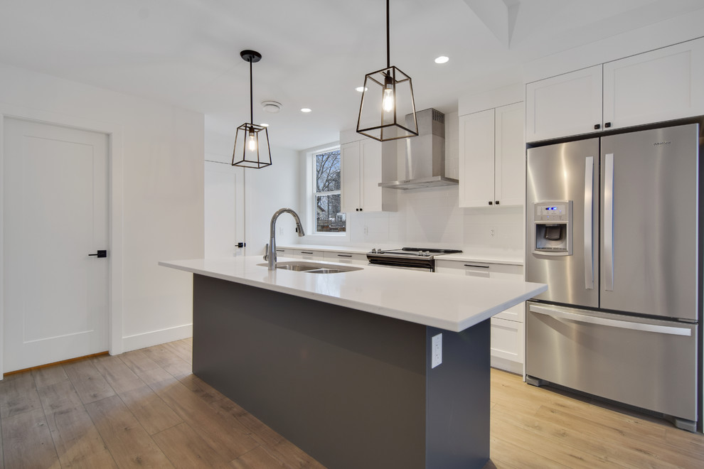 Example of a small trendy galley laminate floor and brown floor open concept kitchen design in Other with an undermount sink, shaker cabinets, white cabinets, solid surface countertops, white backsplash, stainless steel appliances, an island and white countertops