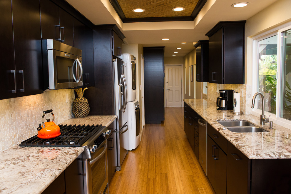 Inspiration for a tropical galley eat-in kitchen remodel in Orange County with an undermount sink, flat-panel cabinets, dark wood cabinets, granite countertops, white backsplash, stone tile backsplash and stainless steel appliances