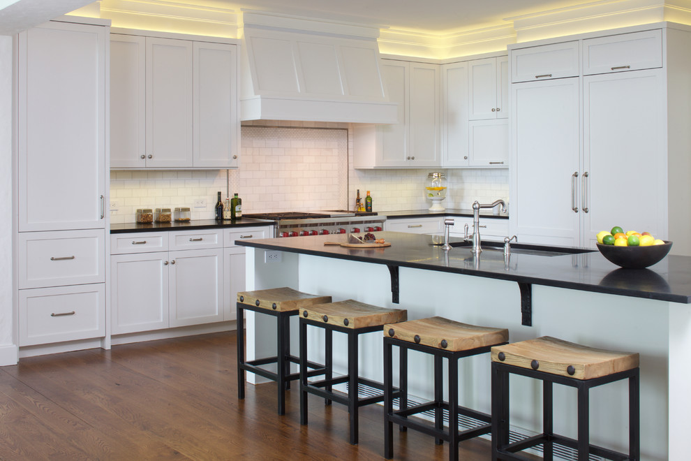 Example of a large classic medium tone wood floor kitchen design in New York with an undermount sink, shaker cabinets, white cabinets, soapstone countertops, white backsplash, subway tile backsplash, paneled appliances and an island