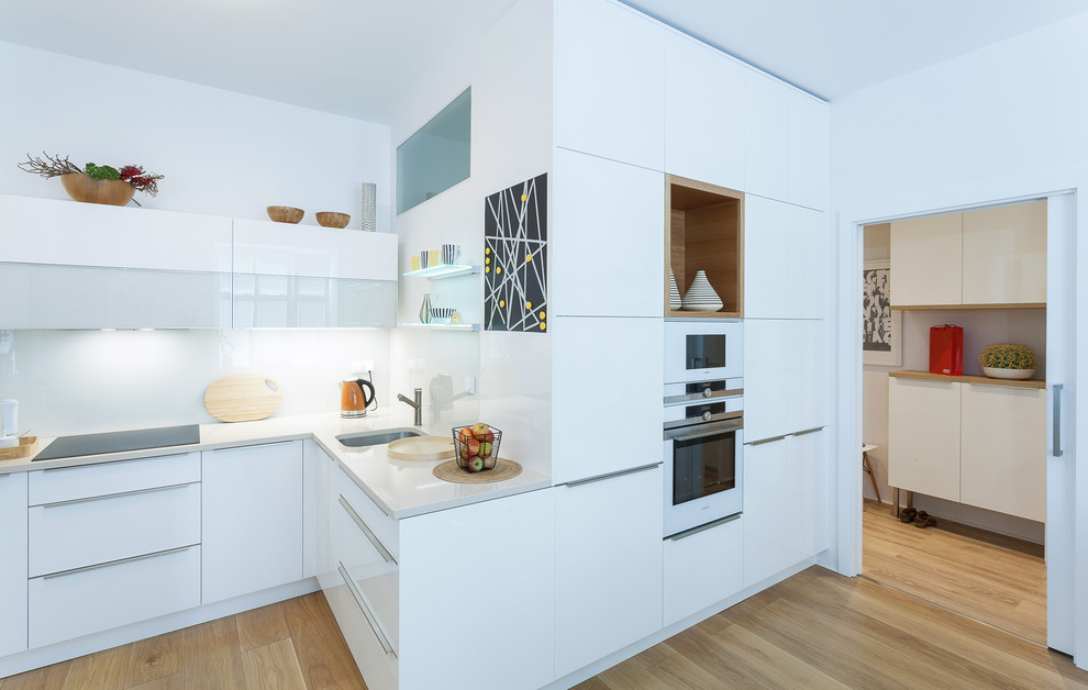 Modern inredning av ett mellanstort kök, med en undermonterad diskho, släta luckor, vita skåp, granitbänkskiva, vitt stänkskydd, glaspanel som stänkskydd, vita vitvaror och ljust trägolv
