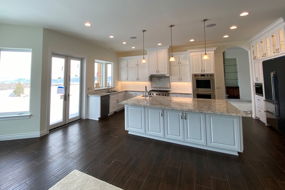 Bild på ett mycket stort funkis grå grått kök, med en undermonterad diskho, luckor med upphöjd panel, vita skåp, granitbänkskiva, vitt stänkskydd, stänkskydd i cementkakel, rostfria vitvaror, klinkergolv i porslin, en köksö och brunt golv