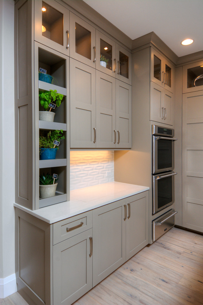 Modelo de cocina actual, grande, con salpicadero blanco, suelo de madera clara, una isla, armarios estilo shaker, puertas de armario grises y electrodomésticos de acero inoxidable