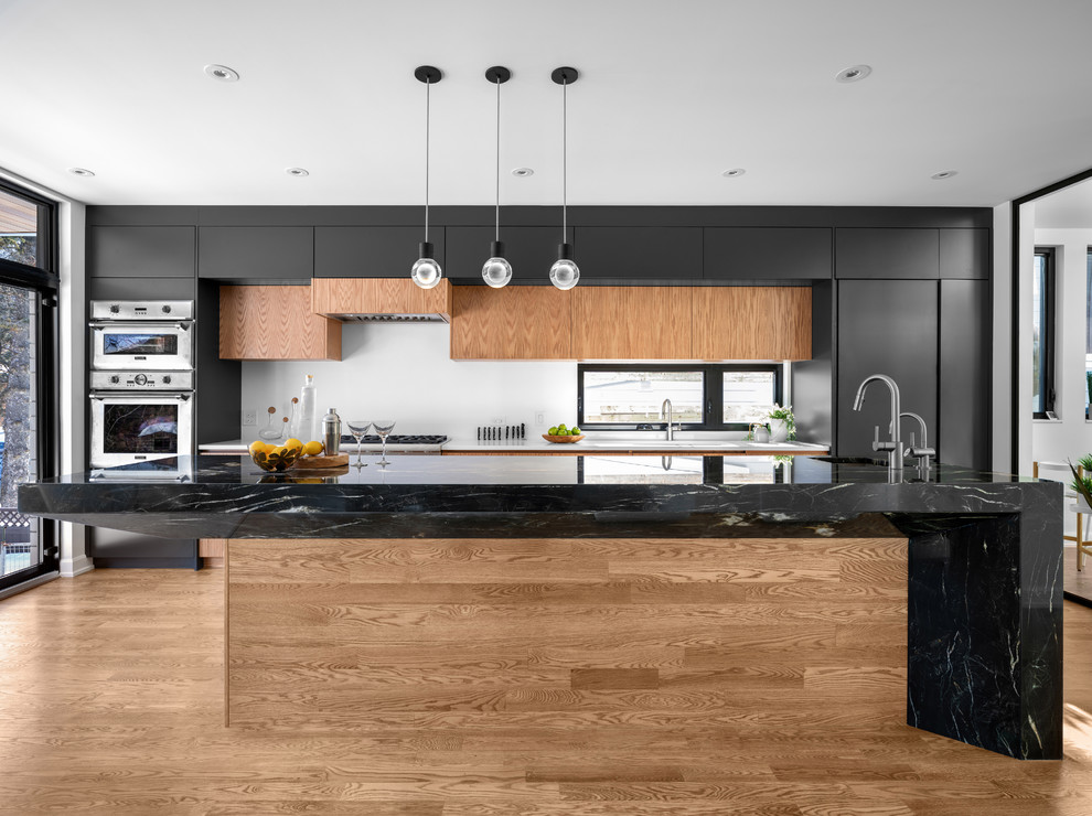 Example of a trendy galley medium tone wood floor and brown floor kitchen design in Ottawa with flat-panel cabinets, black cabinets, white backsplash, stainless steel appliances, an island and black countertops
