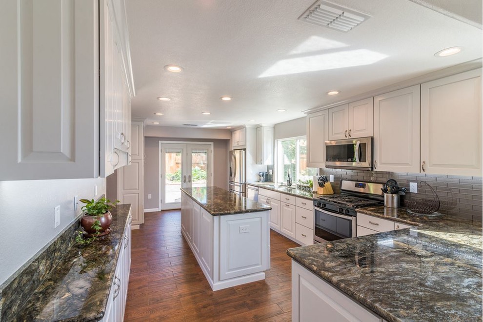 Large contemporary u-shaped kitchen/diner in Sacramento with a submerged sink, raised-panel cabinets, white cabinets, granite worktops, beige splashback, glass tiled splashback, stainless steel appliances, medium hardwood flooring, an island and brown floors.