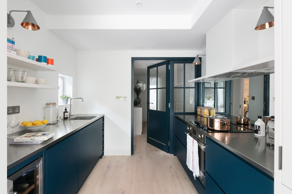 This is an example of a medium sized scandi galley kitchen/diner in London with a single-bowl sink, flat-panel cabinets, blue cabinets, stainless steel worktops, metallic splashback, mirror splashback, integrated appliances, light hardwood flooring and an island.