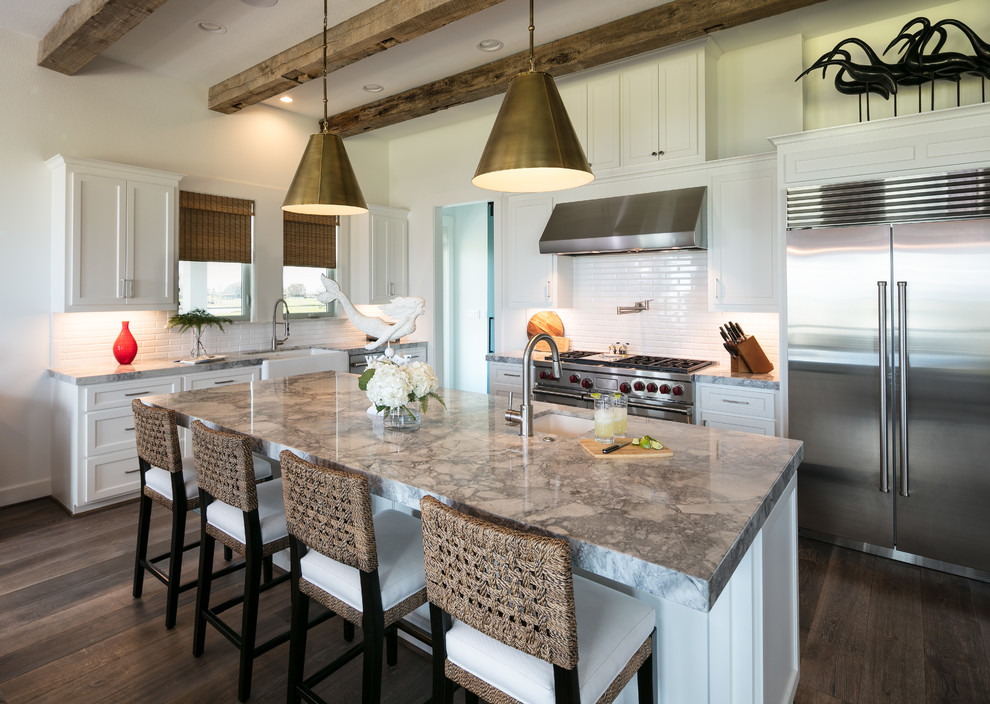This is an example of a beach style l-shaped kitchen in Houston with a belfast sink, shaker cabinets, white cabinets, white splashback, metro tiled splashback, stainless steel appliances, dark hardwood flooring, an island and brown floors.