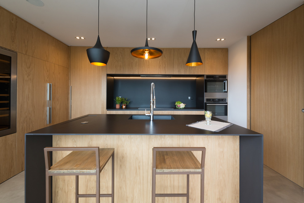 Стильный дизайн: кухня среднего размера в скандинавском стиле с плоскими фасадами, черным фартуком, техникой из нержавеющей стали, островом, светлыми деревянными фасадами, одинарной мойкой и черной столешницей - последний тренд