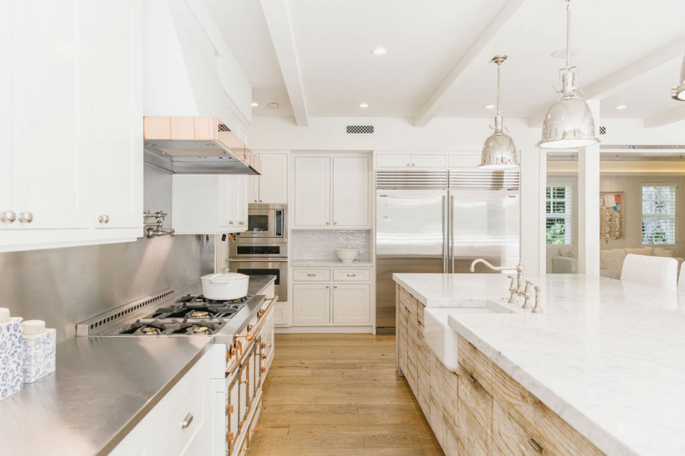 This is an example of a large country u-shaped open plan kitchen in Los Angeles with a belfast sink, recessed-panel cabinets, white cabinets, marble worktops, metallic splashback, porcelain splashback, white appliances, light hardwood flooring, an island, brown floors, white worktops and a wood ceiling.