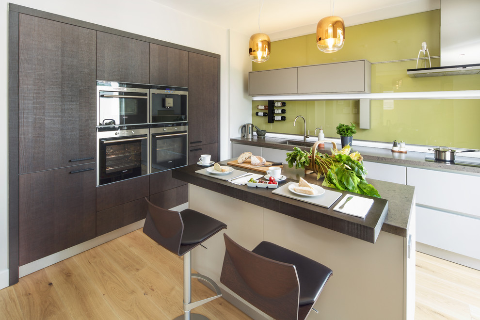 Photo of a medium sized contemporary l-shaped kitchen/diner in Oxfordshire with a single-bowl sink, flat-panel cabinets, dark wood cabinets, engineered stone countertops, green splashback, glass sheet splashback, stainless steel appliances, light hardwood flooring, an island, brown floors and brown worktops.
