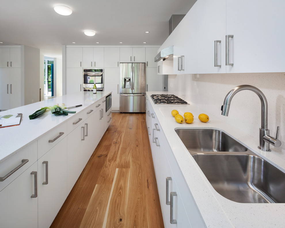 Example of a mid-sized minimalist u-shaped medium tone wood floor and brown floor open concept kitchen design in Portland with a double-bowl sink, flat-panel cabinets, white cabinets, granite countertops, white backsplash, stone tile backsplash, stainless steel appliances, an island and white countertops
