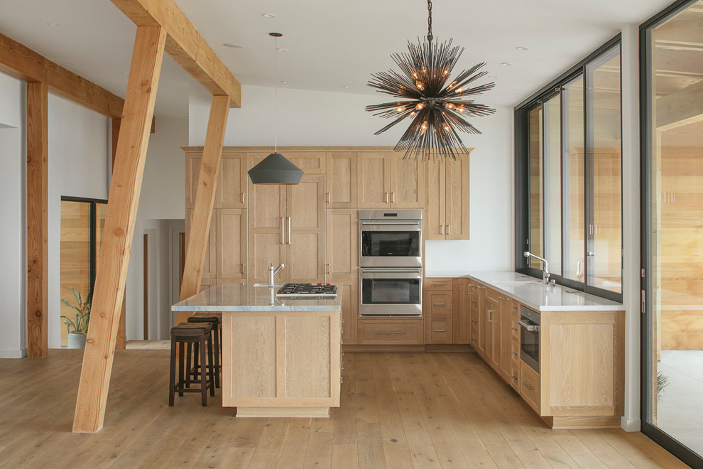 Cette photo montre une cuisine américaine tendance en L et bois clair avec un placard à porte shaker, un électroménager en acier inoxydable, parquet clair, îlot, un sol beige et un plan de travail blanc.