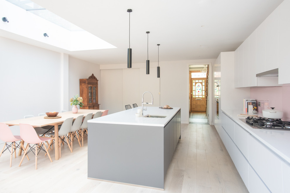 This is an example of a medium sized contemporary single-wall open plan kitchen in London with a built-in sink, flat-panel cabinets, grey cabinets, quartz worktops, pink splashback, glass sheet splashback, light hardwood flooring, an island and white worktops.