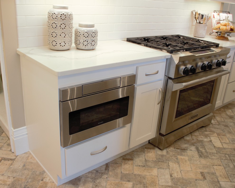 This is an example of a medium sized classic enclosed kitchen in Other with a belfast sink, flat-panel cabinets, white cabinets, engineered stone countertops, white splashback, metro tiled splashback, stainless steel appliances, brick flooring, an island and beige floors.