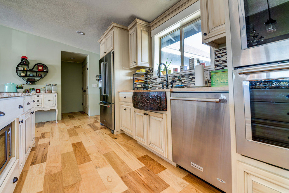 Bild på ett mellanstort lantligt kök, med en rustik diskho, luckor med upphöjd panel, skåp i slitet trä, granitbänkskiva, rostfria vitvaror, ljust trägolv, en köksö, flerfärgad stänkskydd och stänkskydd i mosaik