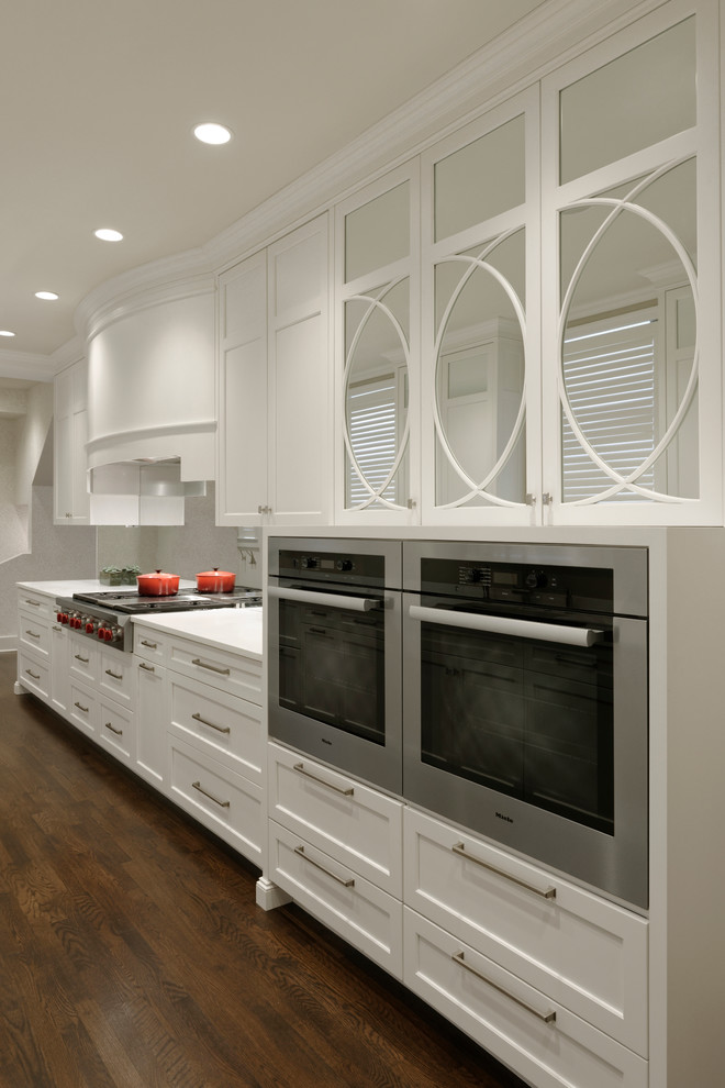 Klassisk inredning av ett mellanstort kök, med en undermonterad diskho, luckor med profilerade fronter, vita skåp, grått stänkskydd, spegel som stänkskydd, rostfria vitvaror, mellanmörkt trägolv och bänkskiva i kvartsit