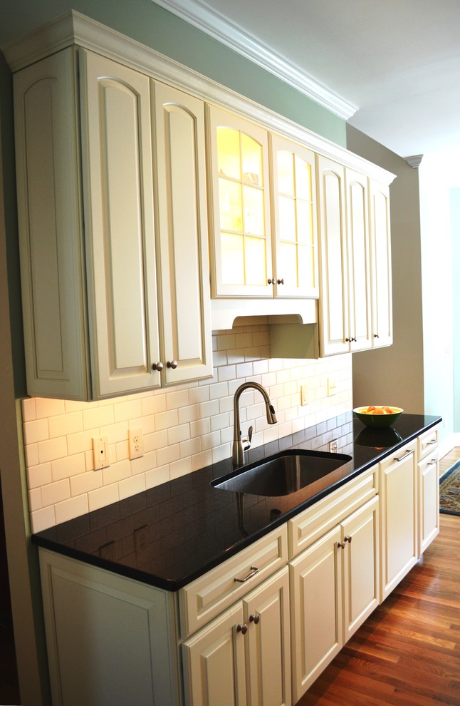 Exempel på ett avskilt klassiskt kök, med en undermonterad diskho, luckor med upphöjd panel, vita skåp, granitbänkskiva, vitt stänkskydd, stänkskydd i tunnelbanekakel, integrerade vitvaror, mellanmörkt trägolv och en köksö