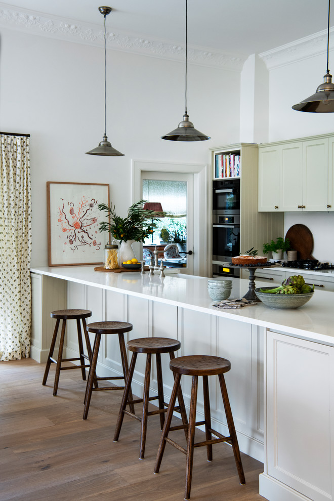 Design ideas for a classic galley kitchen in Sydney with a belfast sink, recessed-panel cabinets, white cabinets, white splashback, black appliances, medium hardwood flooring, a breakfast bar, brown floors and white worktops.