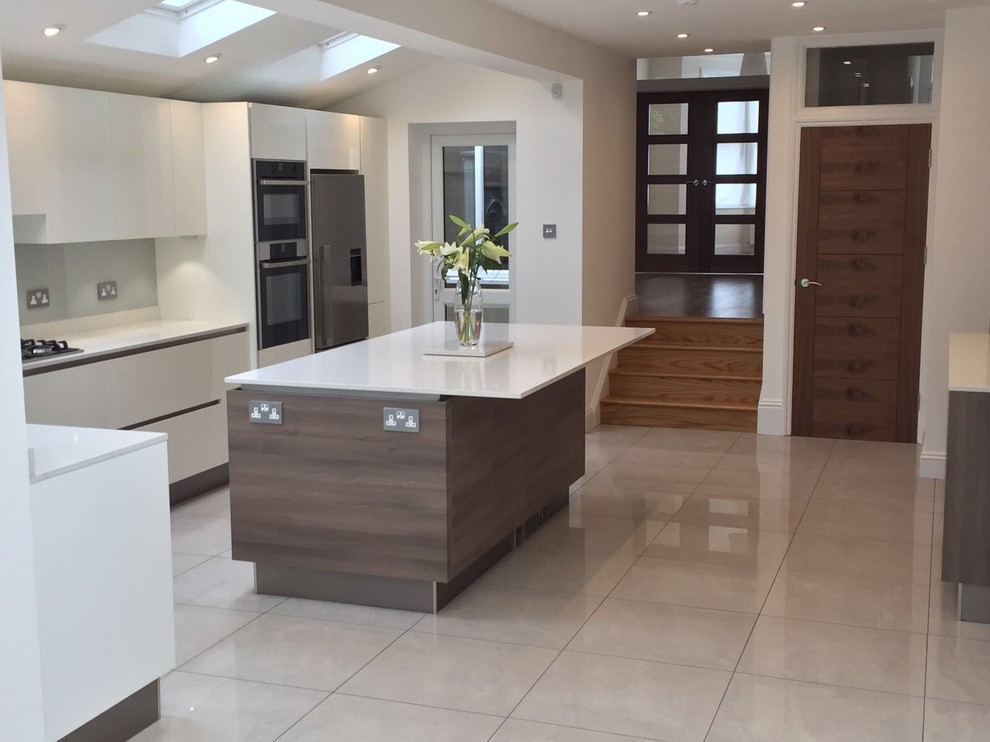 Modern l-shaped kitchen in London with a submerged sink, flat-panel cabinets, white cabinets, composite countertops, metallic splashback, glass sheet splashback, stainless steel appliances, porcelain flooring, an island, grey floors and white worktops.