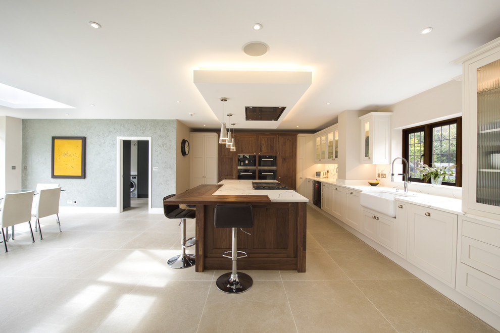 Design ideas for a classic kitchen/diner in Berkshire with a belfast sink, glass-front cabinets, beige cabinets, black appliances, an island, beige floors and white worktops.