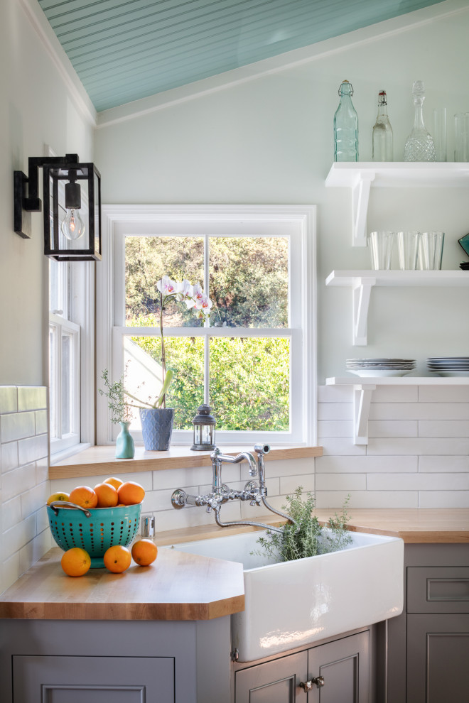 Example of a small beach style galley medium tone wood floor and brown floor enclosed kitchen design in Los Angeles with a farmhouse sink, shaker cabinets, gray cabinets, wood countertops, white backsplash, ceramic backsplash, white appliances, no island and beige countertops