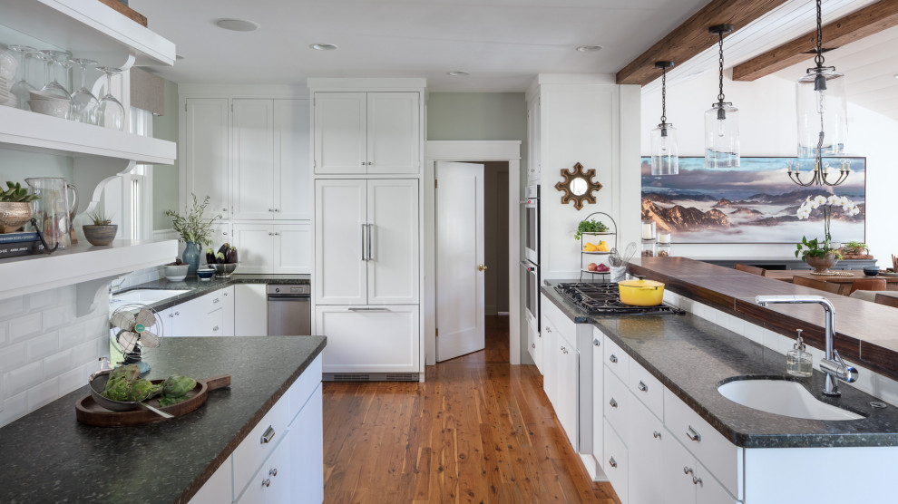 Photo of a medium sized beach style u-shaped enclosed kitchen in Los Angeles with a submerged sink, shaker cabinets, white cabinets, granite worktops, white splashback, metro tiled splashback, integrated appliances, medium hardwood flooring, a breakfast bar, brown floors and black worktops.