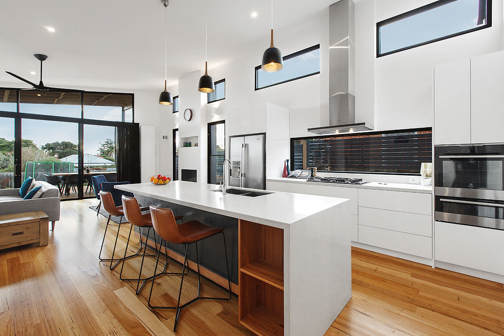 Inspiration for a mid-sized contemporary galley medium tone wood floor open concept kitchen remodel in Melbourne with an undermount sink, flat-panel cabinets, white cabinets, solid surface countertops, white backsplash, ceramic backsplash, stainless steel appliances, an island and white countertops