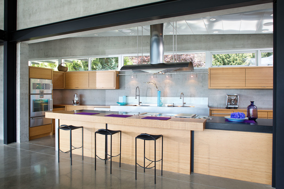 Cette image montre une cuisine ouverte parallèle design en bois clair de taille moyenne avec un placard à porte plane, une crédence grise, un électroménager en acier inoxydable, sol en béton ciré et îlot.