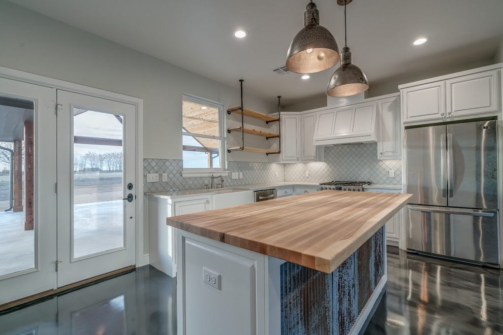 Example of a mid-sized farmhouse l-shaped concrete floor and gray floor open concept kitchen design in Oklahoma City with a farmhouse sink, ceramic backsplash, stainless steel appliances, raised-panel cabinets, white cabinets, wood countertops, white backsplash and an island