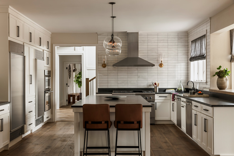 Exemple d'une cuisine chic en L avec un placard à porte shaker, des portes de placard blanches, une crédence blanche, un électroménager en acier inoxydable, un sol en bois brun et îlot.