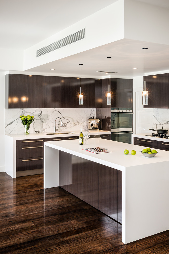 Medium sized contemporary l-shaped open plan kitchen in Melbourne with a submerged sink, flat-panel cabinets, dark wood cabinets, engineered stone countertops, white splashback, stone slab splashback, stainless steel appliances, dark hardwood flooring, an island, brown floors and white worktops.