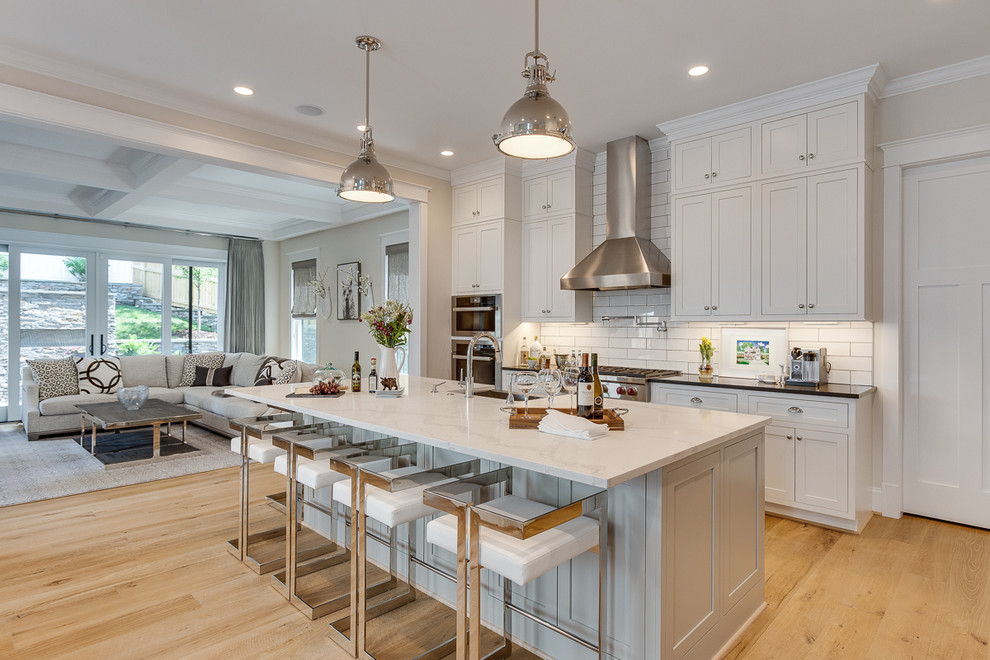 This is an example of a large classic open plan kitchen in DC Metro with shaker cabinets, white splashback, metro tiled splashback, stainless steel appliances, an island, black worktops, a belfast sink, white cabinets, light hardwood flooring, beige floors and granite worktops.