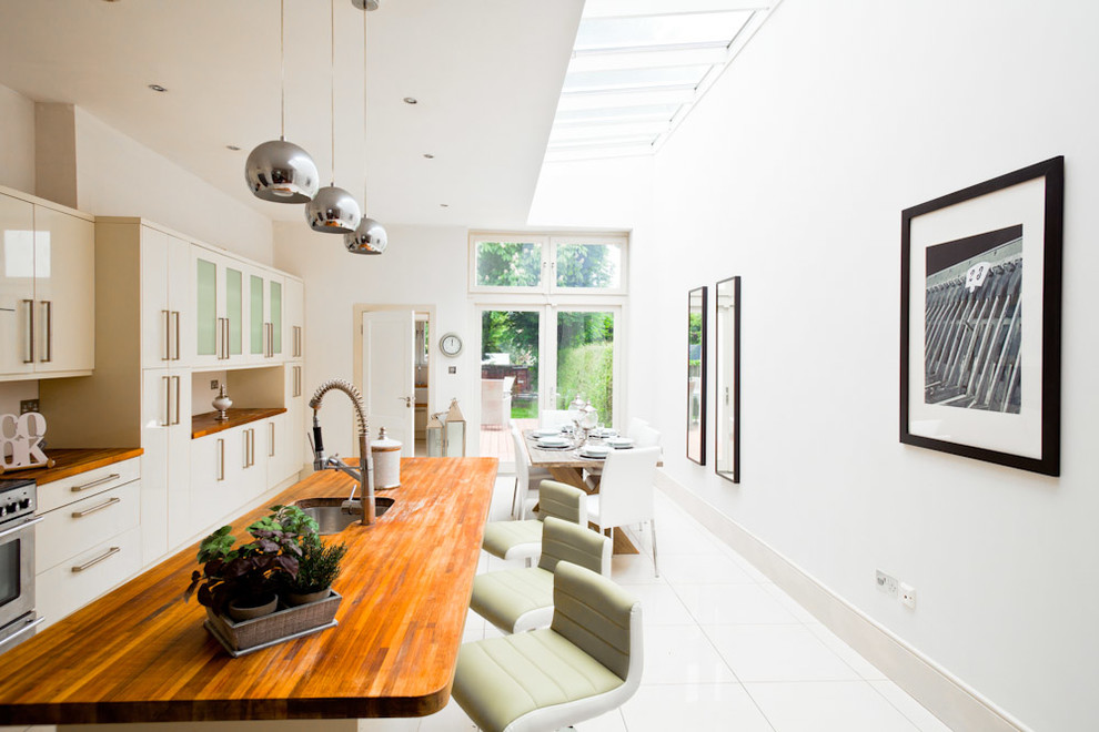 Example of a mid-sized trendy single-wall ceramic tile eat-in kitchen design in Dublin with an undermount sink, flat-panel cabinets, white cabinets, wood countertops, white backsplash, stainless steel appliances and an island