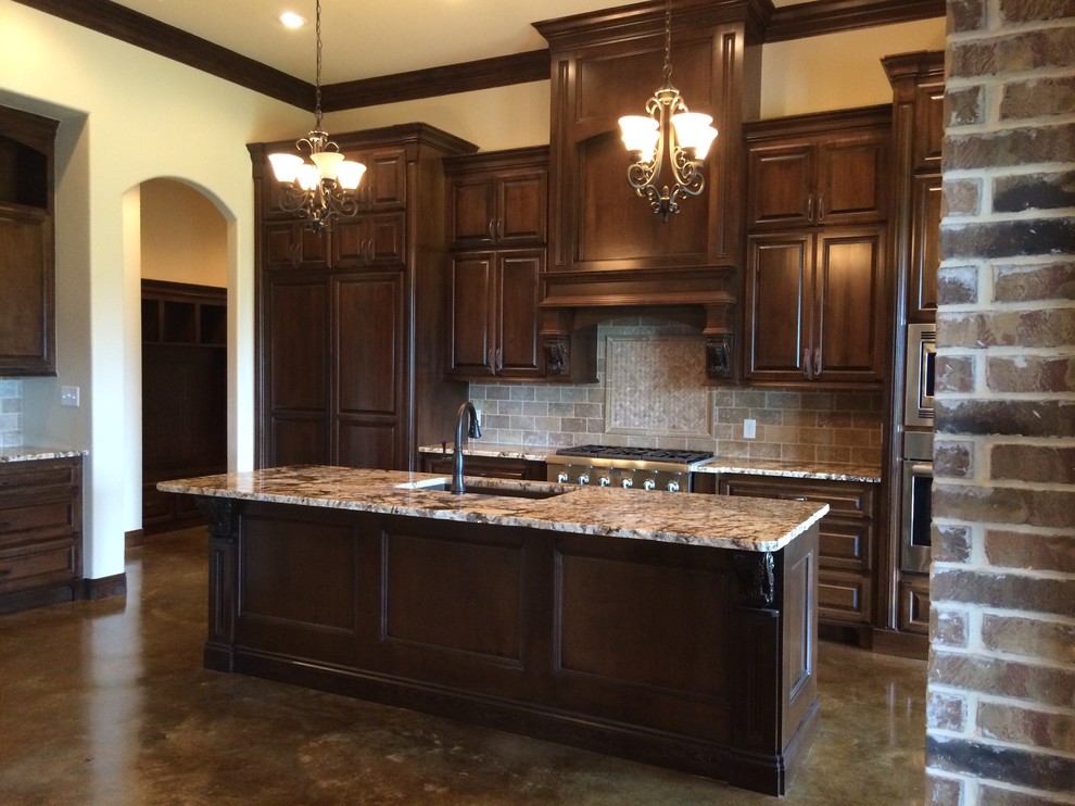 Kitchen - traditional single-wall concrete floor kitchen idea in New Orleans with an undermount sink, raised-panel cabinets, dark wood cabinets, granite countertops, beige backsplash, stone tile backsplash, stainless steel appliances and an island