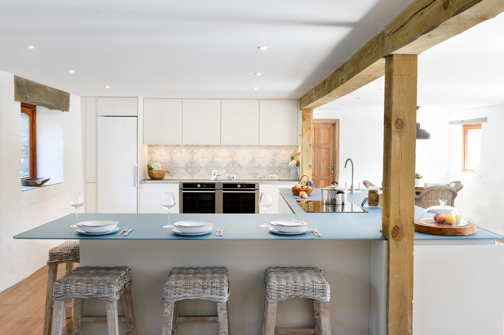 Photo of a large country u-shaped kitchen in Cornwall with a built-in sink, flat-panel cabinets, white cabinets, glass worktops, an island, blue worktops, multi-coloured splashback, stainless steel appliances, medium hardwood flooring and brown floors.