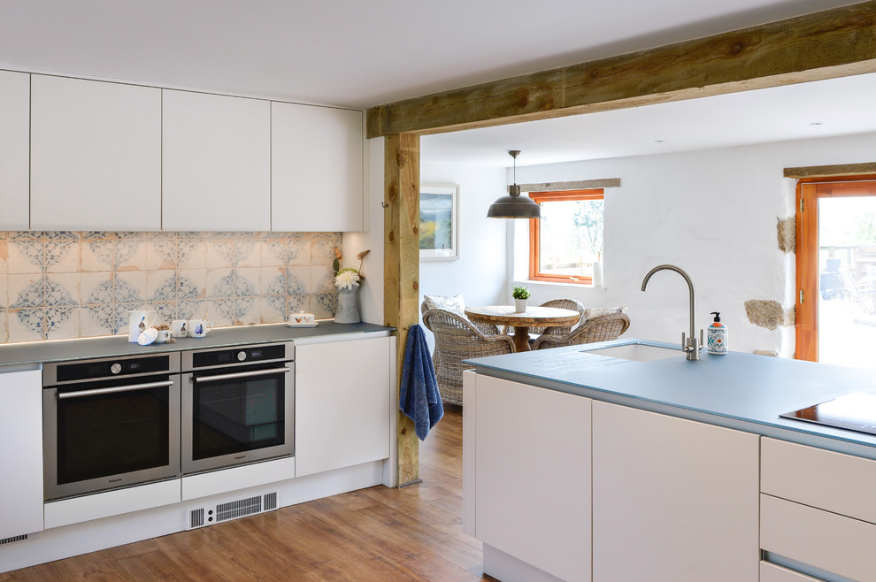 This is an example of a large farmhouse l-shaped open plan kitchen in Cornwall with a built-in sink, flat-panel cabinets, white cabinets, glass worktops, blue splashback, black appliances, an island and blue worktops.
