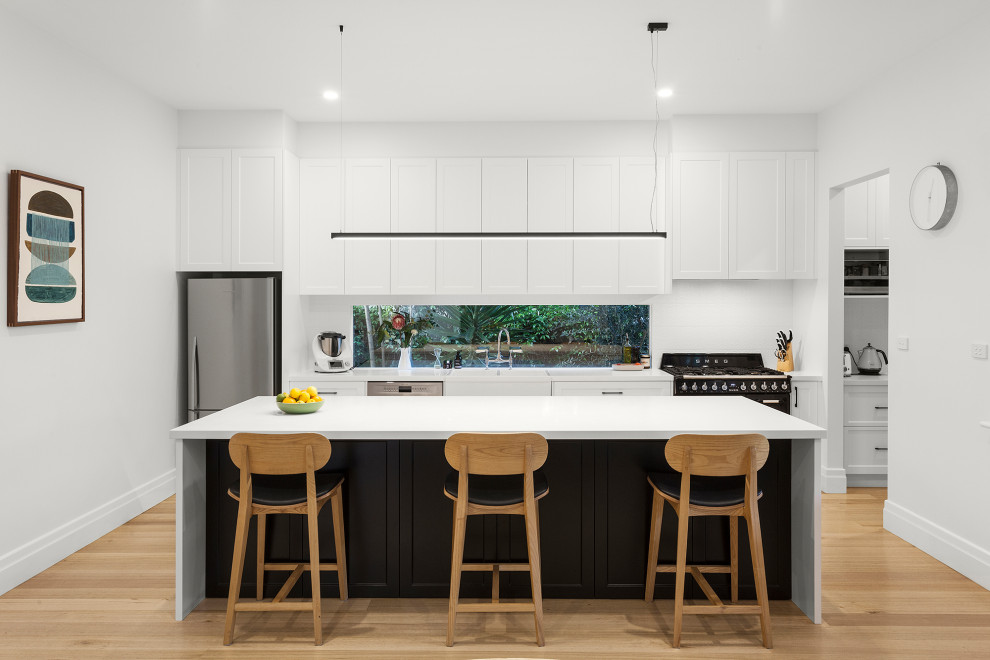 Idéer för mellanstora funkis linjära vitt kök med öppen planlösning, med en rustik diskho, skåp i shakerstil, vita skåp, bänkskiva i kvarts, vitt stänkskydd, fönster som stänkskydd, svarta vitvaror, ljust trägolv, en köksö och beiget golv