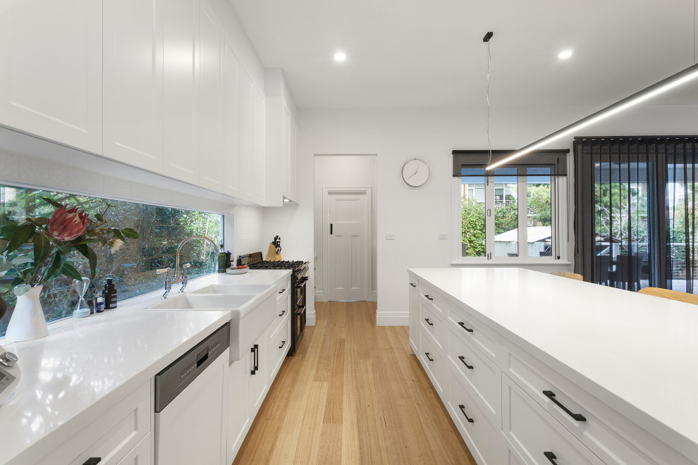 Inspiration for a medium sized contemporary single-wall open plan kitchen in Melbourne with a belfast sink, shaker cabinets, white cabinets, engineered stone countertops, white splashback, window splashback, black appliances, light hardwood flooring, an island, beige floors and white worktops.