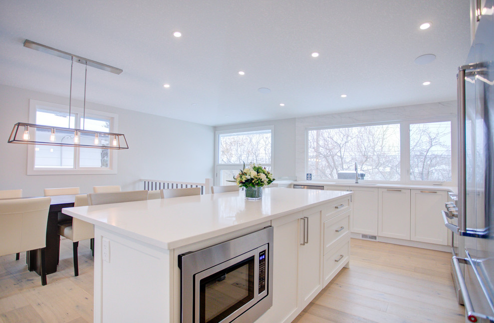 Medium sized contemporary l-shaped open plan kitchen in Calgary with a submerged sink, shaker cabinets, white cabinets, engineered stone countertops, white splashback, ceramic splashback, stainless steel appliances, light hardwood flooring, an island, beige floors and white worktops.