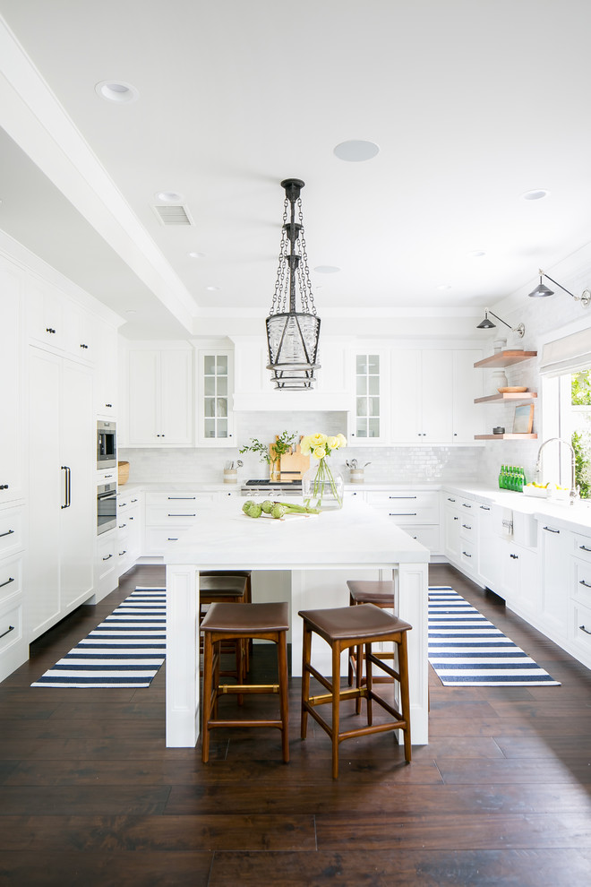 This is an example of a nautical u-shaped kitchen/diner in Orange County with a belfast sink, recessed-panel cabinets, white cabinets, white splashback, stainless steel appliances, dark hardwood flooring, an island, brown floors and white worktops.