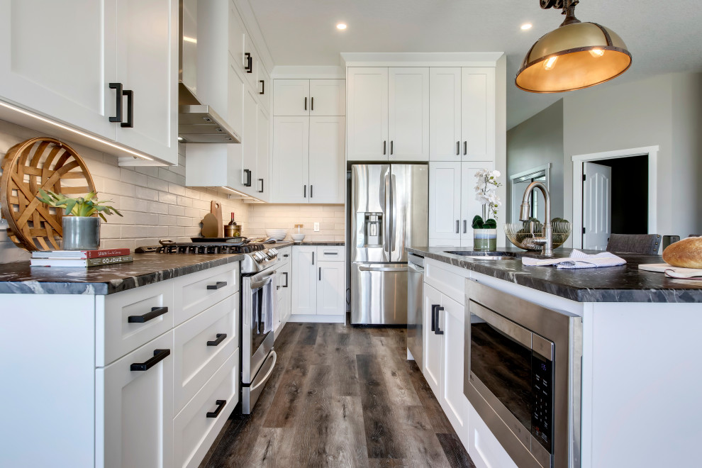 Photo of a medium sized rural l-shaped kitchen in Calgary with a submerged sink, shaker cabinets, white cabinets, granite worktops, white splashback, brick splashback, stainless steel appliances, vinyl flooring, an island, brown floors and black worktops.