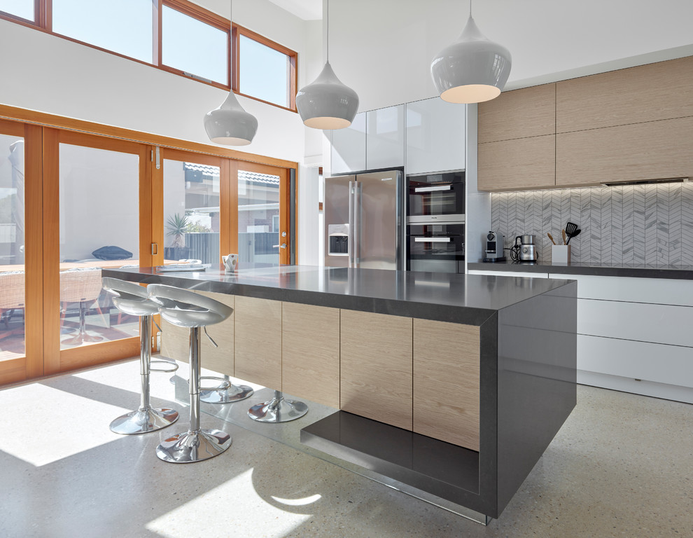 На фото: параллельная кухня среднего размера в современном стиле с плоскими фасадами, техникой из нержавеющей стали, бежевым полом, светлыми деревянными фасадами, островом, белым фартуком и бетонным полом